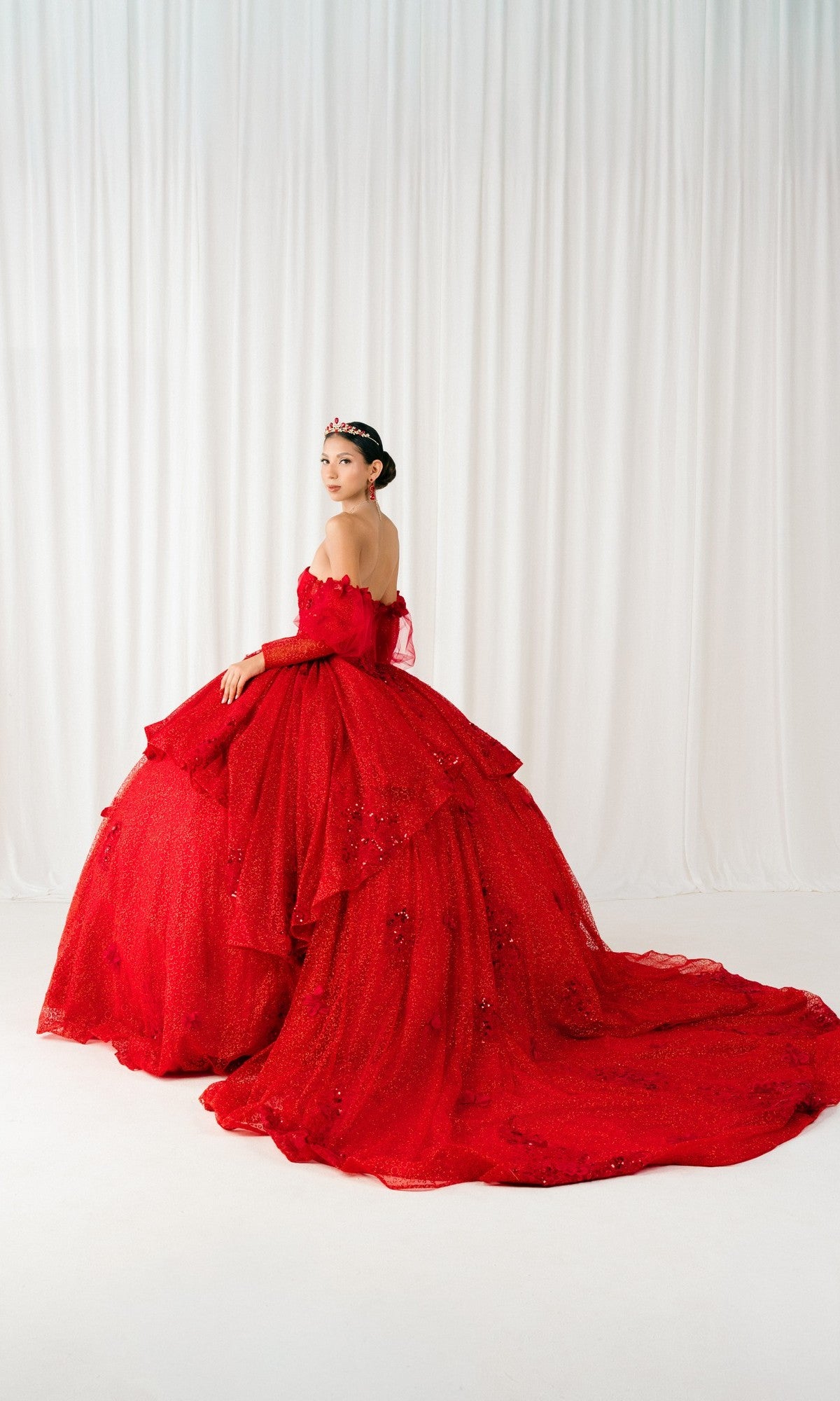 A woman poses before a white curtain in the Quinceanera Dress 8385J By Cinderella Couture—an off-the-shoulder red gown with a corset bodice, long train, and matching red tiara.